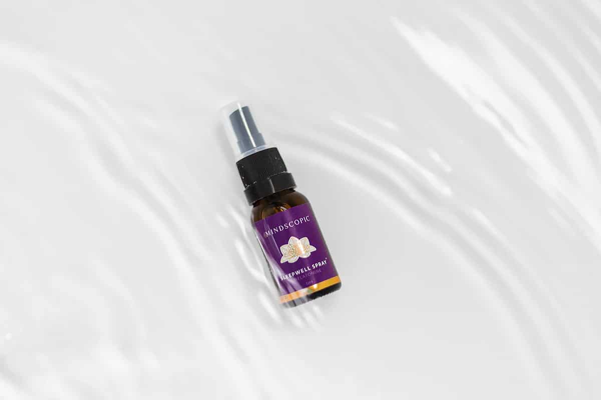 Een klein bruin sprayflesje met het opschrift "MINDSCOPIC - SLEEPWELL SPRAY™ met melatonine (15ml)" met een afbeelding van een vlinder en een paars etiket, geplaatst op een gegolfd wit oppervlak. miniatuur