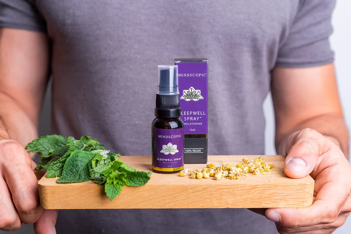 Een persoon houdt een houten plank vast met verse munt, kamille en een flesje MindScopic - SLEEPWELL SPRAY™ met melatonine (15ml) naast het doosje. miniatuur