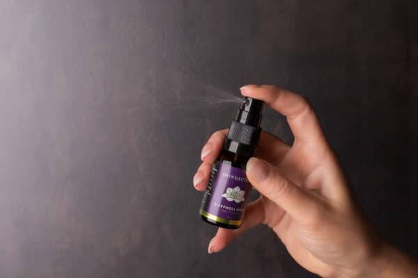 Een hand spuit een klein zwart flesje met de tekst "MindScopic - SLEEPWELL SPRAY™ met melatonine (15ml)" tegen een donkere achtergrond.